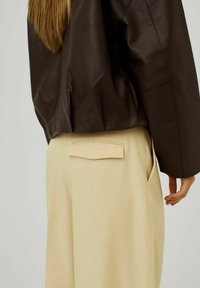 Bruine leren jas met een relaxte pasvorm, gecombineerd met beige wijde broek met een achterzakontwerp. Gelaagde en gladde oppervlakken.