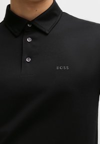 Polo noir avec trois boutons et logo "BOSS" sur la poitrine, porté par une personne montrée de la nuque jusqu'à mi-torse.