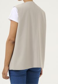 Beige ärmellose Weste mit glatter Textur, gerade geschnittenem Rücken und dezenten Nähten, getragen über einem weißen Hemd und blauen Jeans.