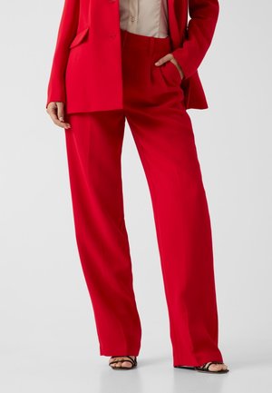Femme portant un pantalon large rouge vif et un blazer assorti avec une chemise beige et des sandales noires à bouts ouverts, main dans la poche.