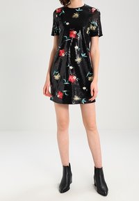 Robe noire pailletée à manches courtes, ornée de broderies florales multicolores. Associée à des bottines noires.