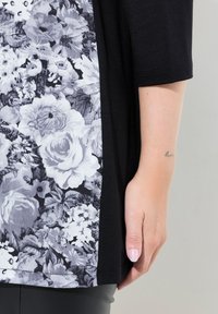 Schwarzes Kleidungsstück mit floralem Muster in Weiß- und Grautönen auf einer Seite, kurzen Ärmeln und einer weichen Textur.