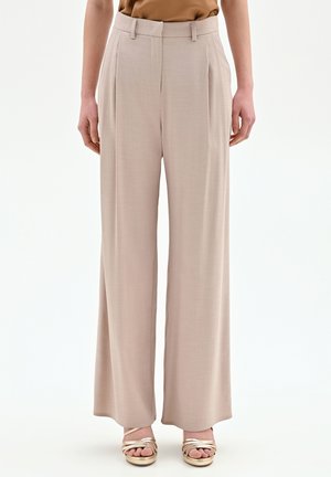 Pantalon beige taille haute à jambes larges, associé à des sandales dorées métalliques à bout ouvert, porté par une personne debout devant un fond uni.