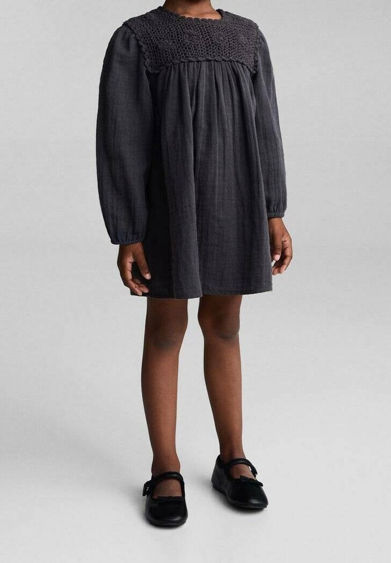 Enfant portant une robe noire à manches longues avec empiècement texturé, debout, les bras détendus le long du corps, chaussée de chaussures Mary Jane noires.
