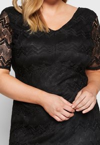 Svart spetsklänning med v-ringad hals och korta, transparenta spetsärmar. Tyget har ett strukturerat, vågigt mönster genom hela.