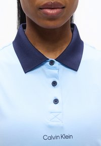 Κοντινό πλάνο ενός ατόμου που φοράει γαλάζιο πόλο πουκάμισο με σκούρο μπλε γιακά και τρία κουμπιά, με το λογότυπο Calvin Klein κάτω από τα κουμπιά.
