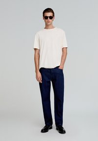 Camiseta de manga corta color crema, jeans rectos oscuros, zapatos negros y gafas de sol negras. Diseño sencillo, tejido de algodón, corte relajado.