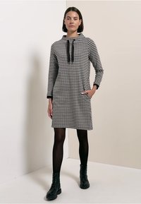 Mustavalkoruudullinen mekko, jossa on putkikaulus, pitkät hihat, sivutaskut ja kiristysnyöri. Yhdistetty mustiin sukkahousuihin ja nauhakenkiin.