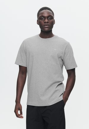 Pilkas trumpomis rankovėmis medvilninis marškinėlis su apvalia iškirpte ir mažu spaudiniu krūtinės srityje. Dėvimi su tamsiomis kelnėmis.