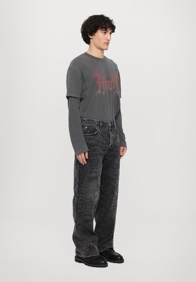 Tricou grafic gri cu un design roșu, suprapus peste o cămașă cu mânecă lungă, asortat cu blugi negri cu model și pantofi negri masivi.