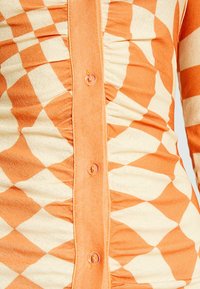 Top in tessuto a quadri arancione e crema con arricciature, dotato di una patta centrale con bottoni trasparenti. Texture morbida.