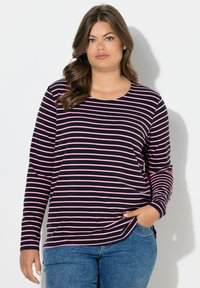 Žena s dlouhými hnědými vlasy, oblečená v černo-růžovém pruhovaném tričku s dlouhým rukávem a modrých džínách, stojící s jednou rukou v kapse.