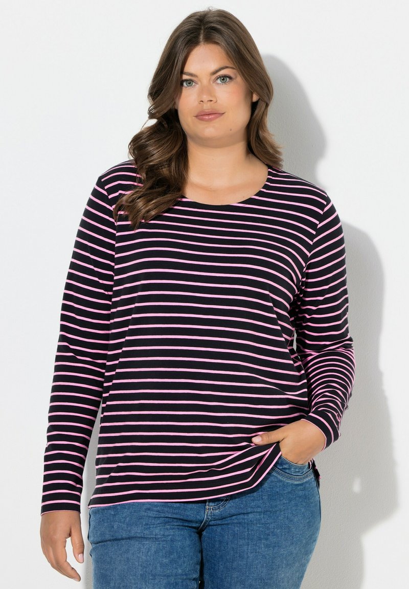 Žena s dlouhými hnědými vlasy, oblečená v černo-růžovém pruhovaném tričku s dlouhým rukávem a modrých džínách, stojící s jednou rukou v kapse.