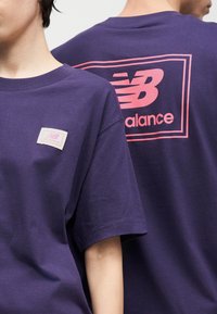 Δύο άτομα με μοβ μπλουζάκια New Balance, με ροζ λογότυπα στο στήθος και μεγάλο λογότυπο στην πλάτη.