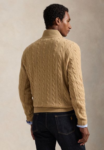 Mand med mørkt krøllet hår iført en beige kabelstrikket sweater og mørke jeans, stående med ryggen og hovedet vendt til siden.