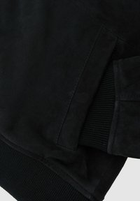 Detail na rukáv černé semišové bundy s žebrovaným úpletem na manžetě a prošitím přední kapsy.