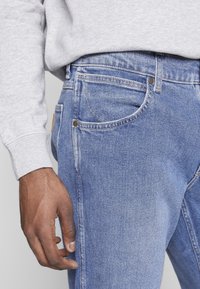 Muž v světle modrých džínech a světle šedém mikině, s levou rukou uvolněně volnou po boku.