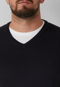 Schwarzer V-Ausschnitt-Pullover aus feinem Strickmaterial, über einem weißen T-Shirt getragen. Dezentes Markenlogo in Hüfthöhe. Saubere gerippte Abschlüsse.