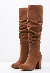 Bottes en suède marron jusqu'au genou avec un design froncé, un talon épais et une texture lisse. Présente un détail de fermeture éclair sur le côté.