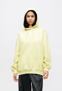 Žltá, nadrozmerná mikina s predným vreckom, kapucňou so šnúrkou a malým bielym logom na hrudi. Textúrovaná zmes bavlny.
