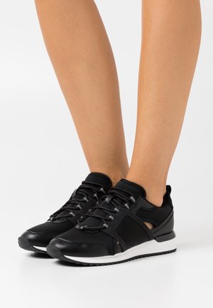Jambes portant des baskets noires à lacets avec des semelles blanches et des panneaux latéraux en mesh sur un fond blanc uni.