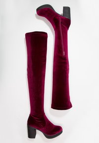 Une paire de bottes en velours bordeaux, hautes, avec des talons épais noirs et des fermetures éclair sur le côté, présentée sur un fond blanc.