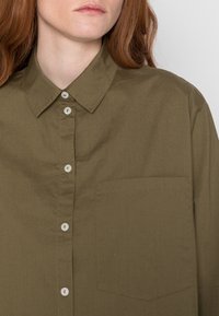 Camisa de botão verde-azeitona feita de algodão, apresentando um colarinho pontudo, botões brancos e um único bolso no peito. Textura suave.
