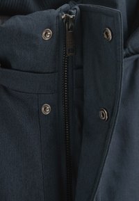 Giacca blu navy in tessuto con finitura texture, dotata di chiusura a zip e bottoni metallici, che mette in evidenza dettagli nella cucitura e nella qualità del materiale.
