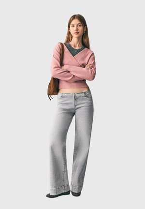 Young woman standing with arms crossed wearing pink sweater, gray wide-leg jeans, black shoes, and brown shoulder bag on gray background.