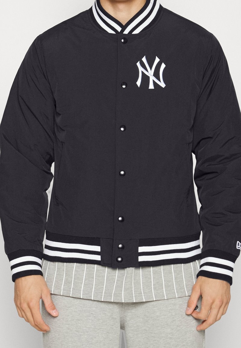 Svart bomberjacka med vita accenter, med en New York Yankees-logotyp, tryckknappar och randiga ribbade manschetter och nederkant.