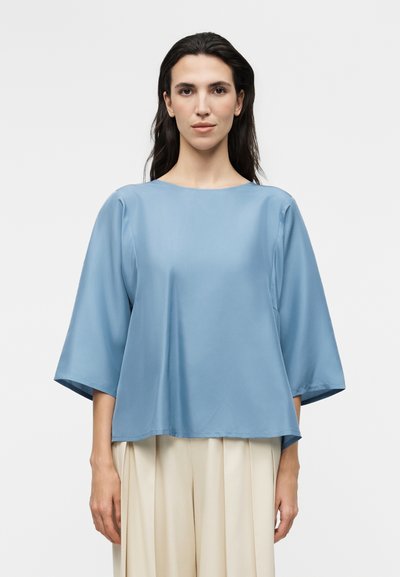 Femme aux cheveux raides et foncés porte un chemisier ample bleu clair à larges manches et un pantalon plissé crème, debout devant un fond uni.