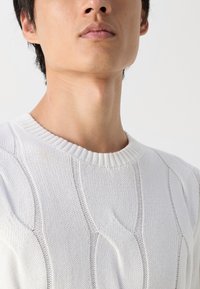 Valkoinen neulepaita, jossa on teksturoitu aaltoilu kuvio, ribattu kaula-aukko ja kevyttä materiaalia. Suunnittelussa keskitytään pystysuoriin viivoihin.