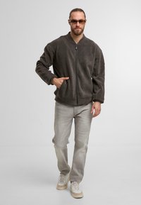 Bomber jacket in pile grigio scuro con zip frontale, tasche laterali e polsini elasticizzati, abbinato a jeans grigio chiaro e sneakers color crema.