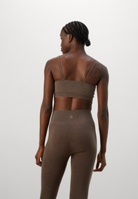 Mulher com cabelo trançado, vestindo um top cropped canelado castanho a condizer e leggings de cintura alta, de pé e de costas contra um fundo simples.