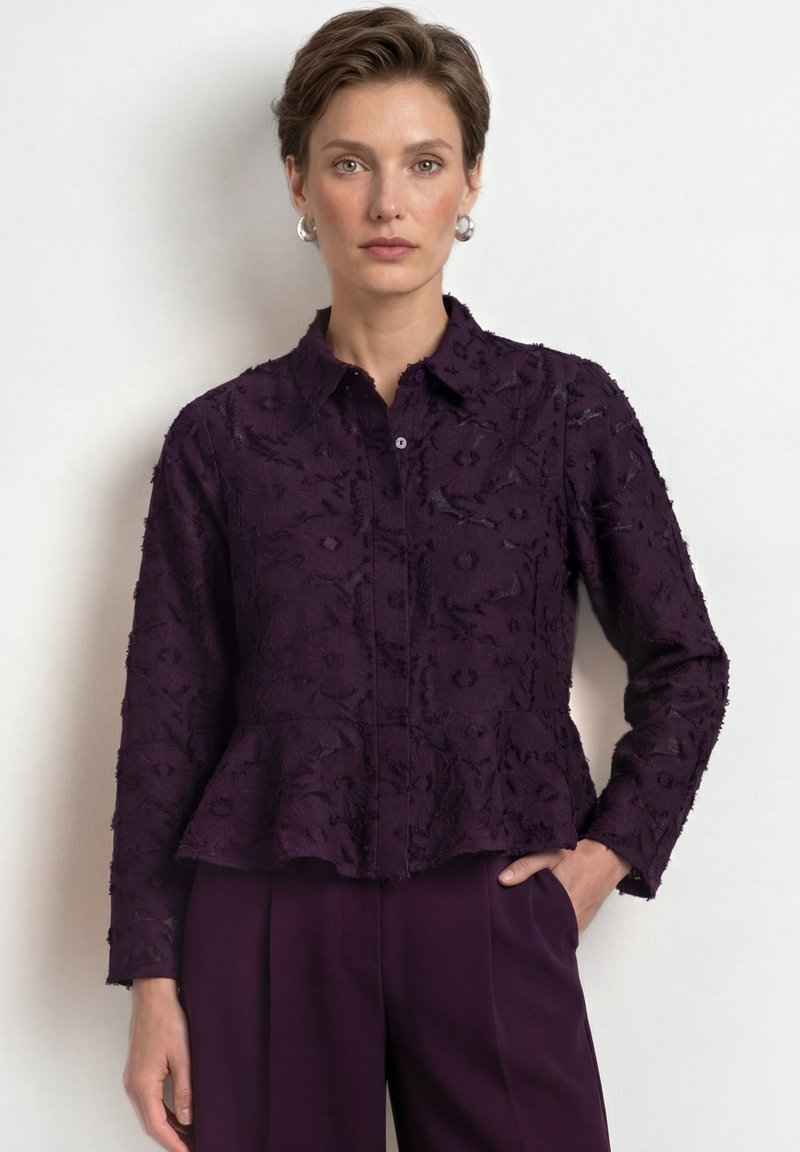 Woman with short hair wearing a textured dark purple buttoned blouse and matching trousers, standing against a plain light background.