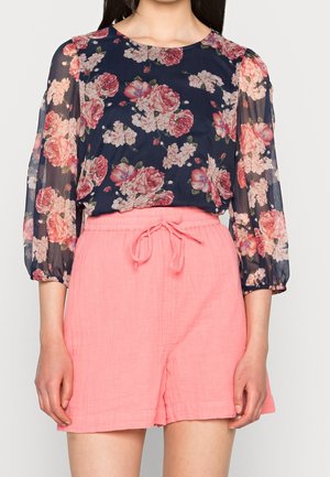 Femme portant un chemisier transparent bleu marine à fleurs avec manches longues et un short rose à cordon, sur un fond uni.