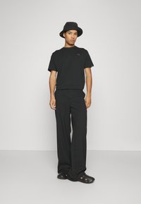 Black t-shirt with short sleeves, black wide-leg pants, and black bucket hat. Shoes are black clogs with cut-out details. Minimalist design.