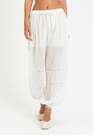 Donna che indossa pantaloni harem larghi, bianchi e semi-trasparenti con pannelli in pizzo e polsini elastici, abbinati a scarpe bianche intrecciate.