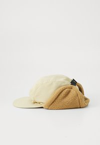 Beige cap with a flat brim, featuring a soft, tan fleece lining and a black adjustable strap at the back. Textured fabric contrast evident.