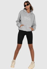 Grauer Kapuzenpullover mit grafischem Design, kombiniert mit schwarzen Leggings und beigen Sneakers. Glatte Textur, lockere Passform, Fronttasche.
