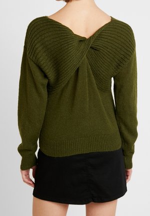 Olive green knitted sweater with twisted ribbed detail on the back, worn over a black mini skirt, shown from behind.