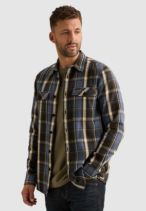 Homme portant une chemise à carreaux bleue, noire et jaune par-dessus un t-shirt vert olive, avec une main dans la poche, regardant sur le côté.