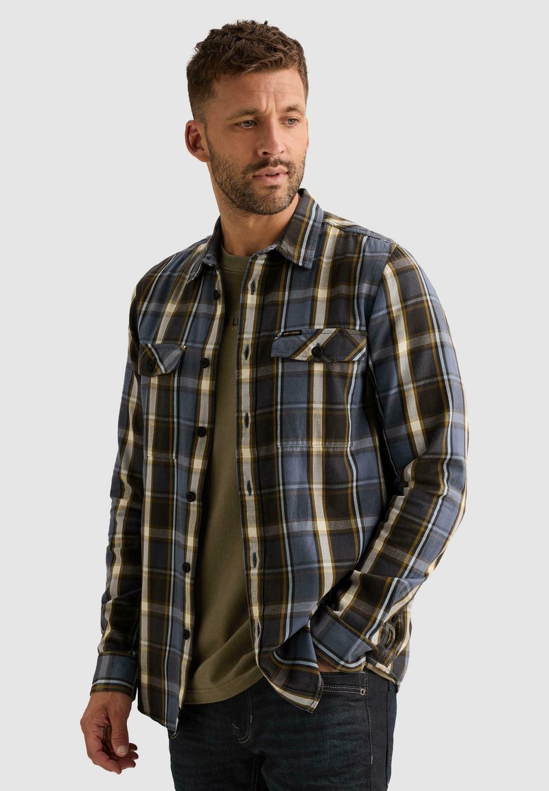 Homme portant une chemise à carreaux bleue, noire et jaune par-dessus un t-shirt vert olive, avec une main dans la poche, regardant sur le côté.