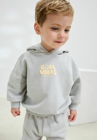 Hellgrauer Kapuzenpullover mit einem lächelnden Gesicht und dem Schriftzug "COOL VIBES" in Creme. Dazu passende Sweatpants.