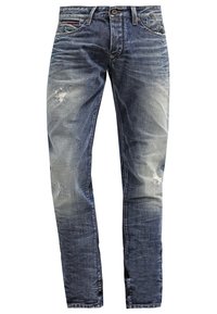 Tummansiniset farkut kuluneilla yksityiskohdilla ja haalistumilla. Niissä on perinteinen viiden taskun malli sekä vetoketjullinen haara, jossa on napillinen suljenta.