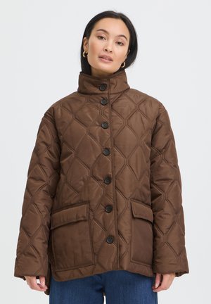 Femme portant une veste matelassée marron avec de gros boutons et deux poches à rabat à l'avant, associée à un jean bleu, sur un fond blanc uni.