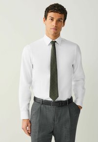 Biela košeľa s zelenou kravatu s bodkami. Model nosí pruhované sivé nohavice a čierny opasok, čo vytvára profesionálny vzhľad.