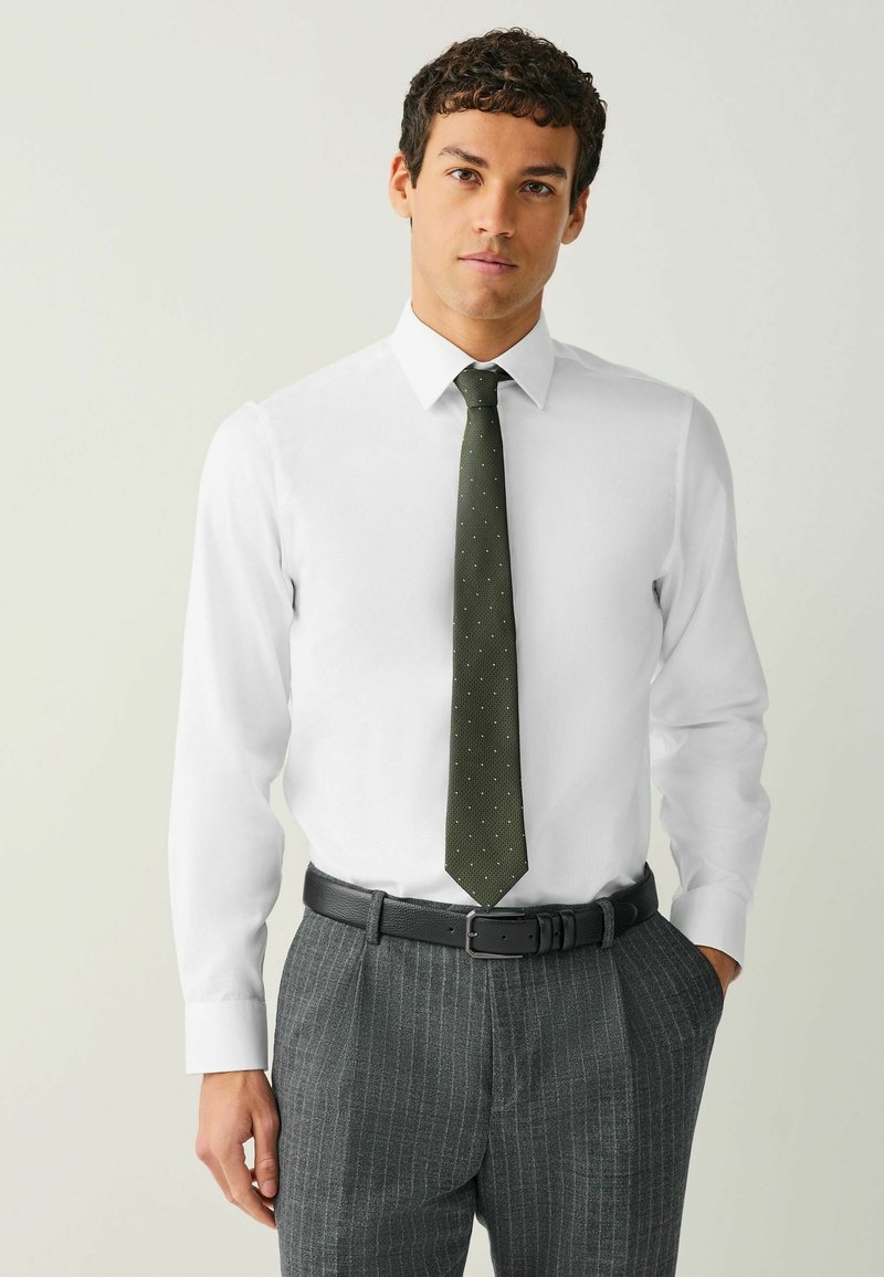 Biela košeľa s zelenou kravatu s bodkami. Model nosí pruhované sivé nohavice a čierny opasok, čo vytvára profesionálny vzhľad.