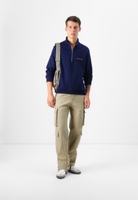 Un modello maschile è in piedi contro uno sfondo bianco, indossando un maglione navy con zip e pantaloni cargo beige, mentre tiene una borsa a tracolla grigia.