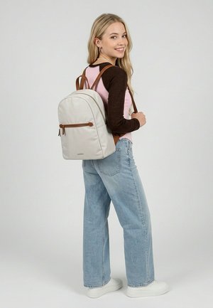 Jauna moteris dėvi šviesiai mėlynas džinsus, baltus sportinius batelius, rausvą ir rudą ilgomis rankovėmis palaidinę, šypsosi su smėlio spalvos kuprine ant nugaros, stovi šonu.